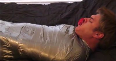 A young man lies bound tightly in silver duct tape mummy bondage on a bed while wearing a red ball gag.