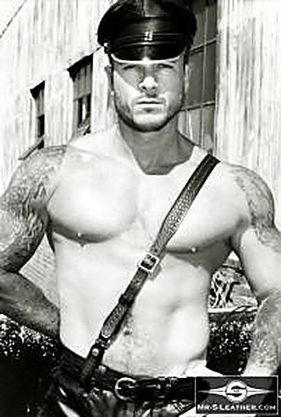 A muscular, tattooed man wears a black leather Muir cap and cross-chest leather strap outdoors against a warehouse background.