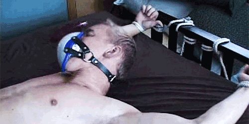 A mature man lies restrained on a bed with white rope tied to his wrists and a leather harness mask over his face.