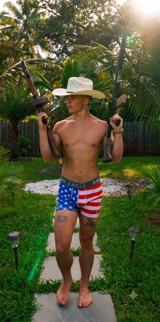 A shirtless muscular man wearing American flag boxer briefs and a cowboy hat holds two rifles while standing on a garden path.
