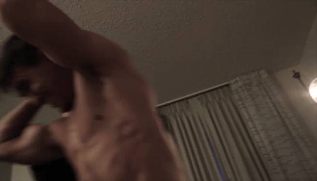 A shirtless muscular man with defined abdominal muscles moves within a dimly lit bedroom setting.