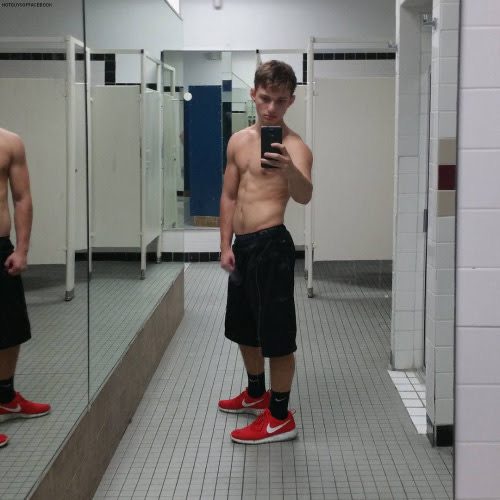 A shirtless twunk in black gym shorts and red sneakers takes a mirror selfie in a tiled locker room.