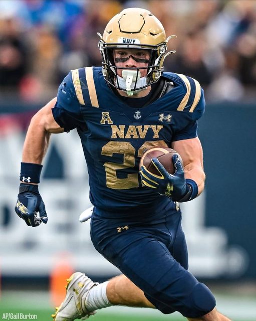 A muscular American football player in a Navy uniform runs with the ball on the field during a game.