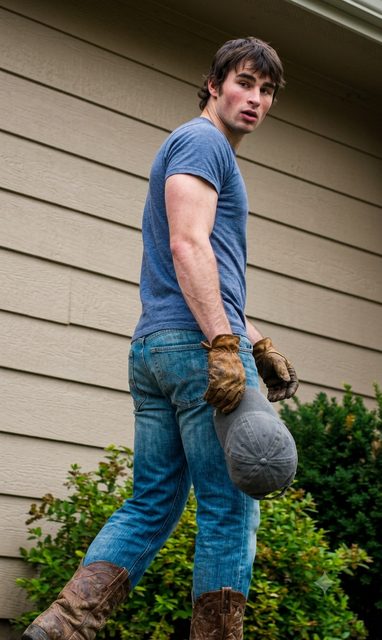 A muscular young man in a blue t-shirt, denim jeans, and leather cowboy boots looks back while wearing work gloves and holding a cap outdoors.