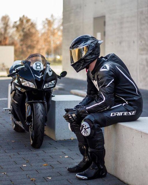 A man in a full black leather Dainese racing suit and a camouflage helmet sits beside his sport motorcycle.