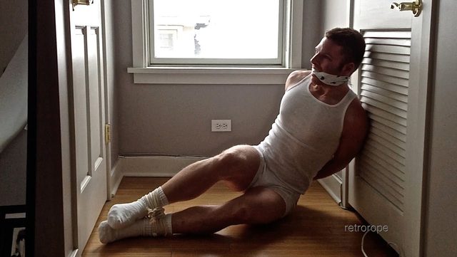 A muscular man wearing white underwear and socks sits on a wooden floor with his hands tied behind his back, his ankles bound with rope, and a cloth gag in his mouth.
