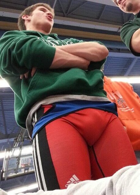 A low-angle perspective shows a young man wearing a green hoodie over a tight red and blue Adidas wrestling singlet.