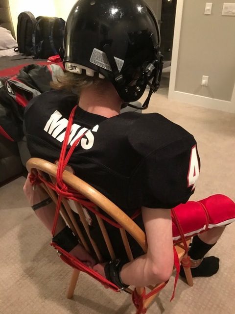 A man in a football uniform and helmet is tied to a wooden chair with red rope and black leather cuffs.