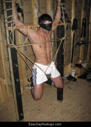 A muscular man wearing white gym shorts and a blindfold is suspended in an intricate rope bondage setup in an unfinished basement.