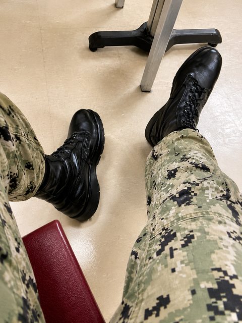 A first-person perspective shows a person wearing digital green camouflage trousers and polished black leather combat boots while sitting.