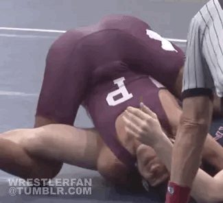 A wrestler wearing a maroon singlet pins his opponent to the mat during a grappling match.