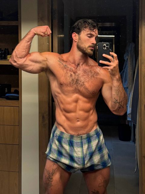 A muscular, bearded man with a hairy chest and tattoos takes a mirror selfie while flexing his bicep in plaid boxer shorts.