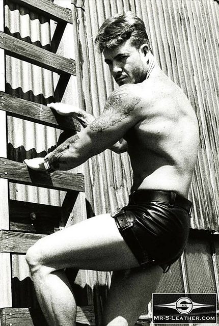 A muscular tattooed man wearing tight black leather shorts climbs a wooden ladder against a corrugated metal wall.