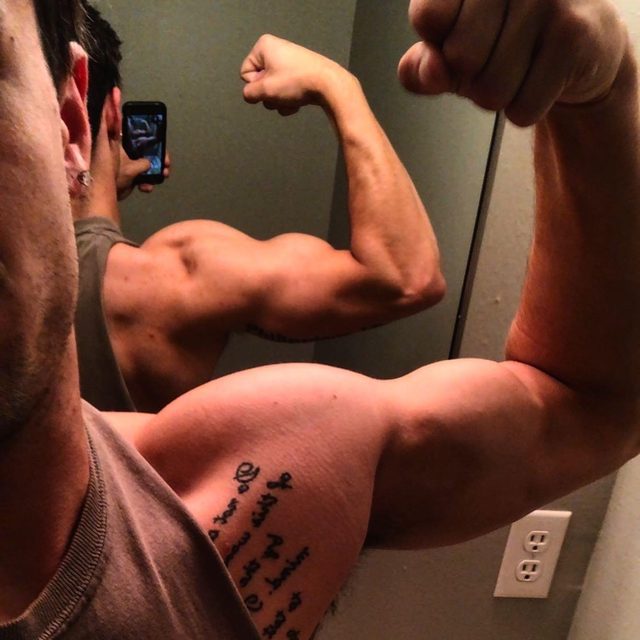 A muscular man captures a mirror selfie while flexing his large bicep, showcasing a tattoo on his inner arm.