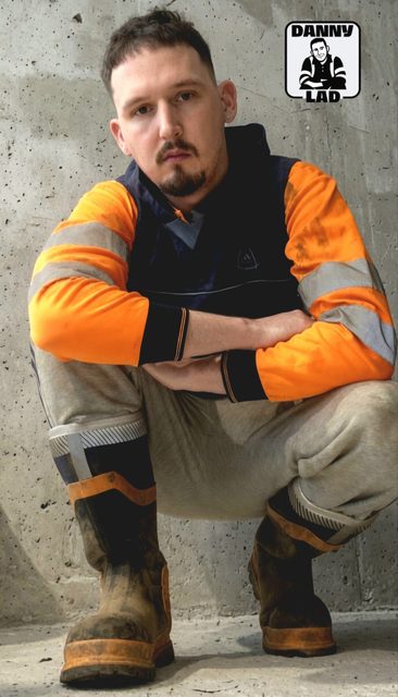 A bearded man in a high-visibility shirt and grey sweatpants squats in front of a concrete wall while wearing muddy work boots.