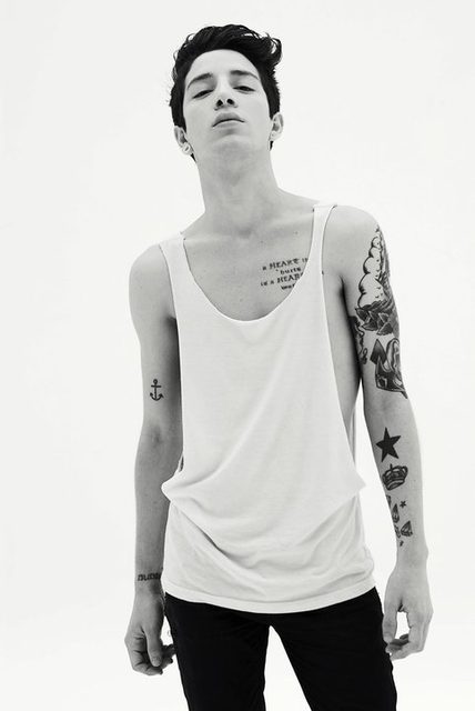 A slim, tattooed twink poses in a deep-cut white tank top and black jeans in a black and white studio shot.