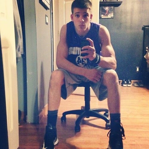 A muscular young man in a sports jersey and gym shorts takes a mirror selfie while sitting on an office chair.