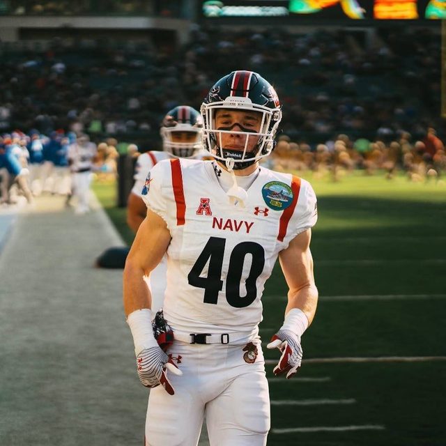 A muscular young man in a full Navy football uniform stands on a stadium field.