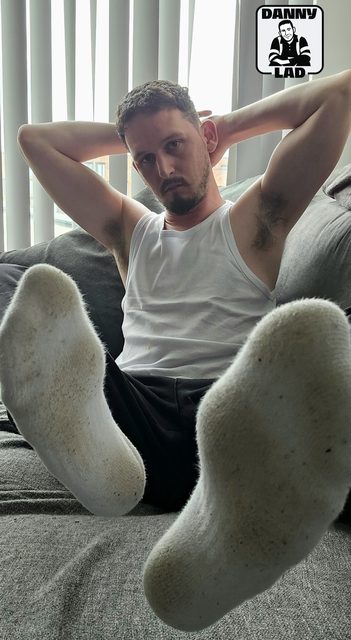 A bearded man in a white tank top sits on a couch with his hands behind his head, prominently displaying the soles of his dirty white socks to the camera.