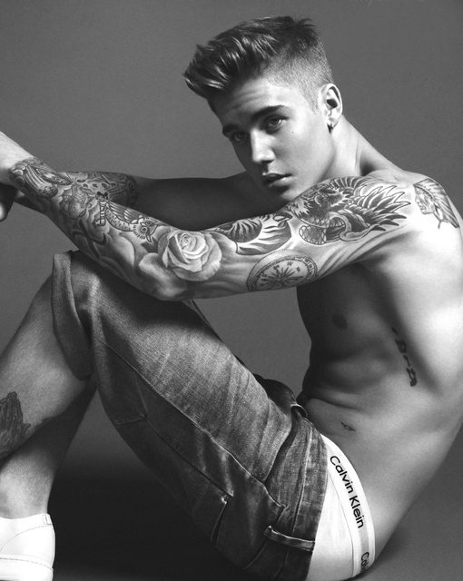 A shirtless twunk with extensive tattoos on his arms and torso poses in cuffed denim shorts and white underwear.