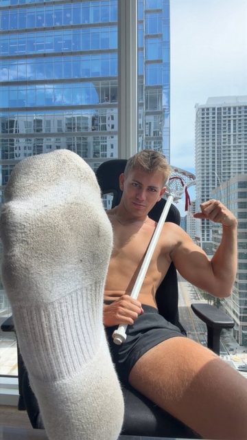 A muscular man in socks and shorts holds a lacrosse stick in a high-rise city apartment.