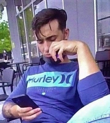 A young man with tattoos sits at an outdoor cafe, holding a small bottle of poppers to his nose while looking at his smartphone.