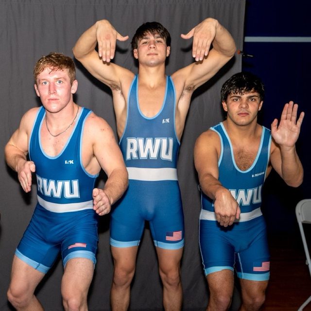 Three young men in blue wrestling singlets pose for a group photo indoors.