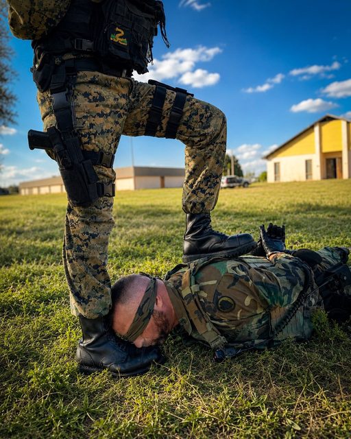 A man in a military uniform lies submissively on the grass while another soldier in tactical gear presses a combat boot onto his back and holds another near his face.