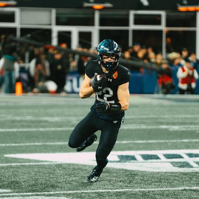 A muscular football player in a black uniform and helmet runs across the field carrying the ball.