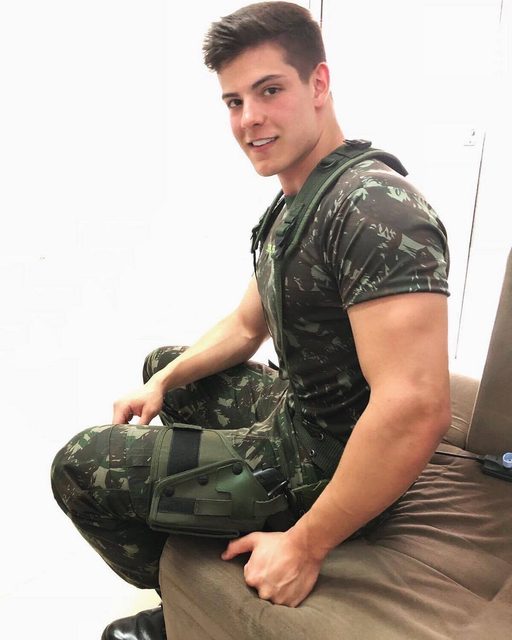 A muscular young man in a full military camouflage uniform and tactical gear sits on a couch.
