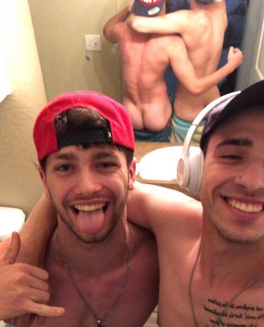 Two shirtless young men pose for a playful bathroom selfie while one man's bare buttocks are visible in the mirror reflection behind them.