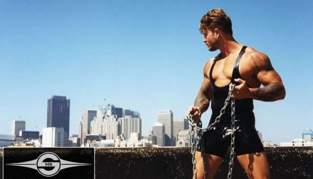A muscular tattooed man stands on a rooftop wearing a black rubber singlet while holding heavy metal chains against a city skyline.