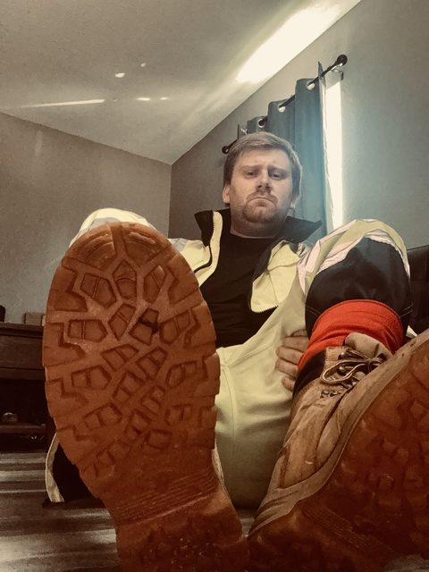 A bearded man in a high-visibility construction uniform sits on the floor, presenting the soles of his work boots in a low-angle POV shot.