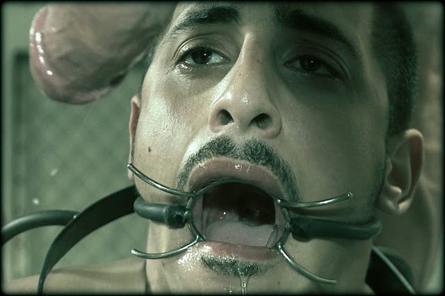 A submissive man wearing a metal mouth gag looks up as an erect penis hangs near his face.