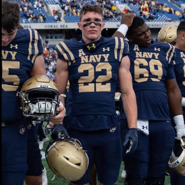 A group of muscular football players in full navy and gold uniforms stand on the field, with the central player wearing eye black and holding his helmet.