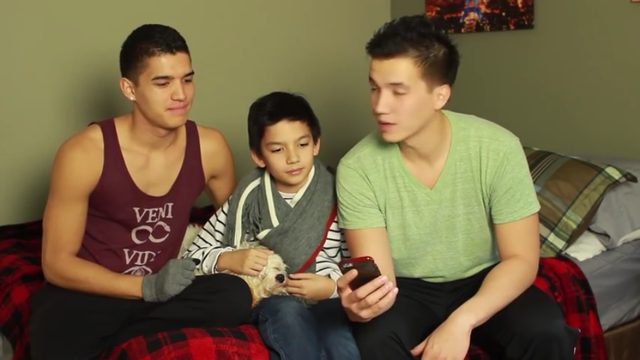 Two young men and a child sit on a bed with a small dog while looking at a phone.