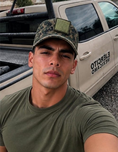 A muscular man in an olive green t-shirt and digital camouflage cap stands outdoors in front of a tan military truck.