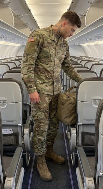 A tattooed man in a US Army camouflage uniform stands in the aisle of an airplane cabin while holding a duffel bag.