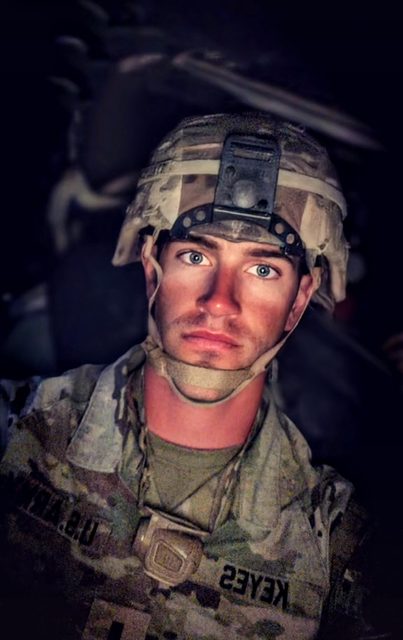 A young man with blue eyes and light stubble wears a full military camouflage uniform and tactical helmet.