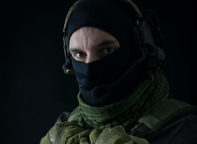 A man in tactical gear poses with a black balaclava, green shemagh, and communication headset against a dark background.