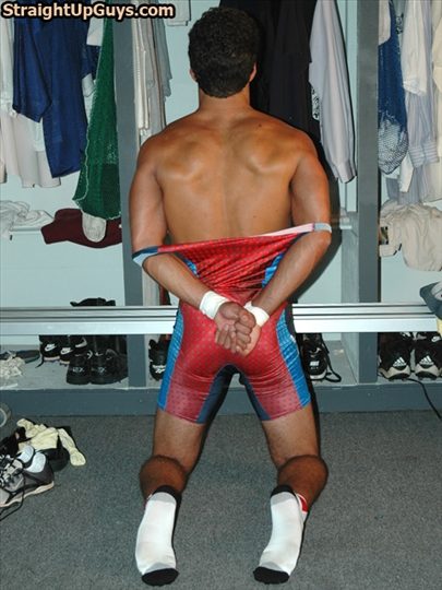 A muscular man kneels in a locker room with his wrestling singlet pulled down and hands behind his back.