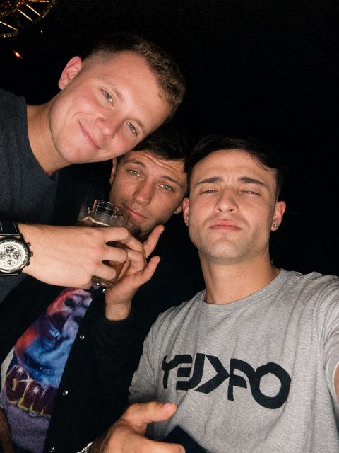 Three young men pose for a close-up selfie in a dimly lit bar or club setting.