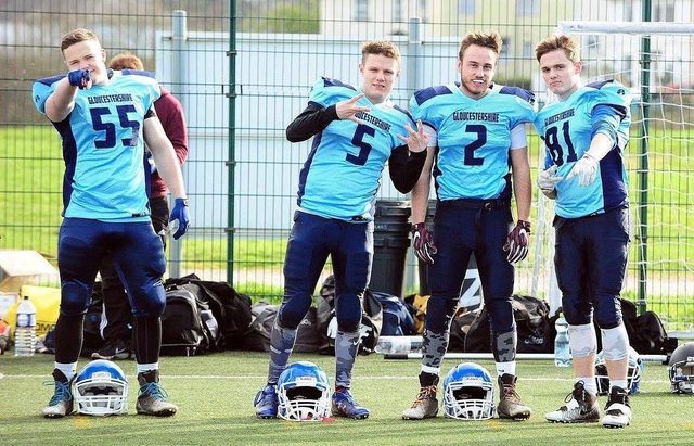 Four young men pose on an outdoor sports field wearing light blue and dark blue American football kits.