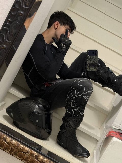A young man in a black compression top, motorcycle boots, and gloves poses for a mirror selfie on a staircase next to his helmet.
