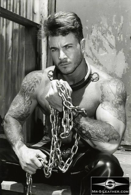 A shirtless, tattooed muscular man poses with heavy metal chains and a metal collar while wearing leather pants.