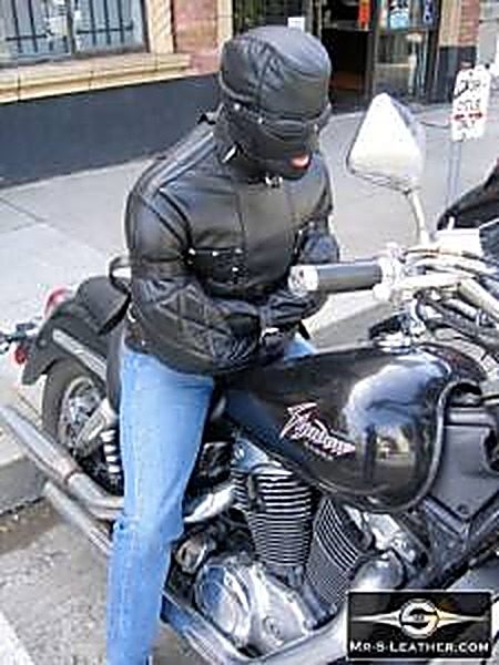 A person wearing a black leather straitjacket and a matching full leather hood sits bound atop a motorcycle on a city sidewalk.