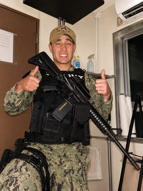A man in a full military camouflage uniform and tactical gear holds a rifle while giving a thumbs up.