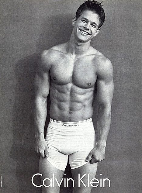 A black and white studio portrait of a muscular, smooth-chested man wearing white Calvin Klein boxer briefs.