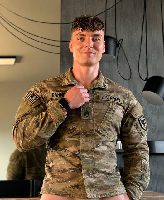 A fit young man in a U.S. Army camouflage uniform jacket poses indoors, looking directly at the camera while adjusting his collar.