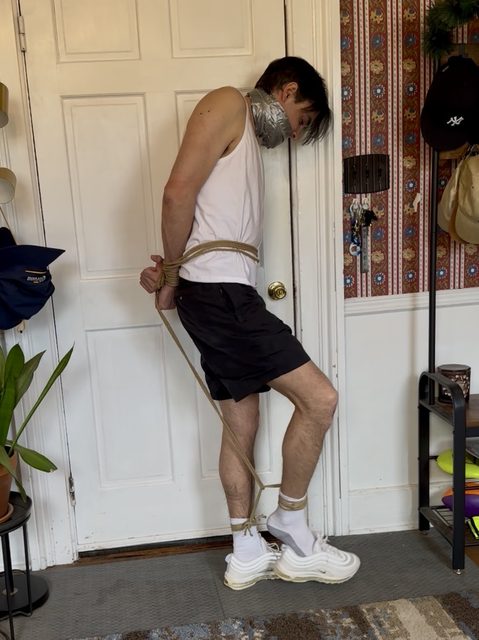 A slim man in a white tank top and black shorts is gagged with duct tape and bound with rope at his waist, wrists, and ankles.